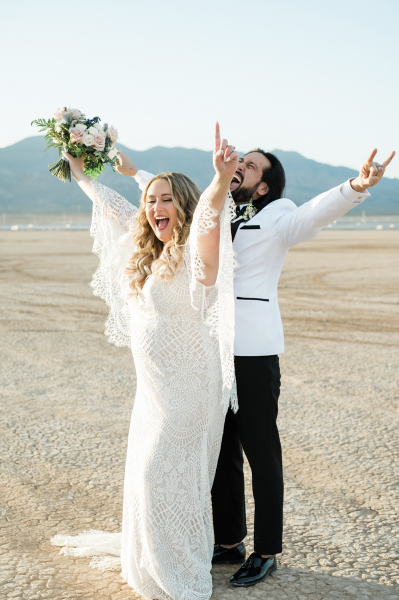 A couple celebrates their wedding on the Dry Lake Bed in Boulder City, Nevada. They stand together with the man behind the woman and each raise their arms and scream with excitement. A couple celebrates their wedding on the Dry Lake Bed in Boulder City, Nevada. They stand together with the man behind the woman and each raise their arms and scream with excitement.