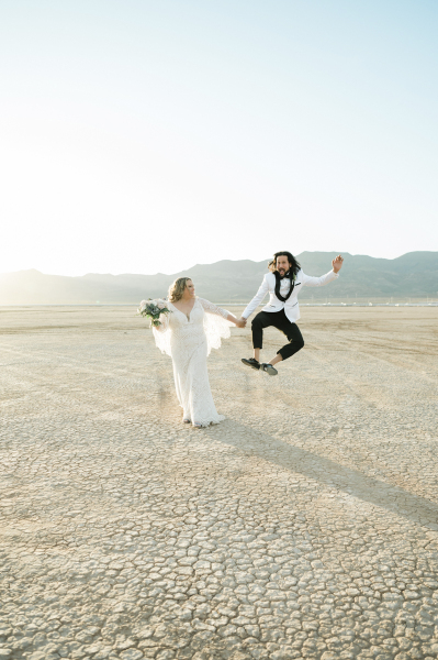 A bride holds hands with a groom while the groom jumps into the air and clicks his heels.The photo is filled with the dry cracked ground of the Dry Lake Bed south of Las Vegas, Nevada.