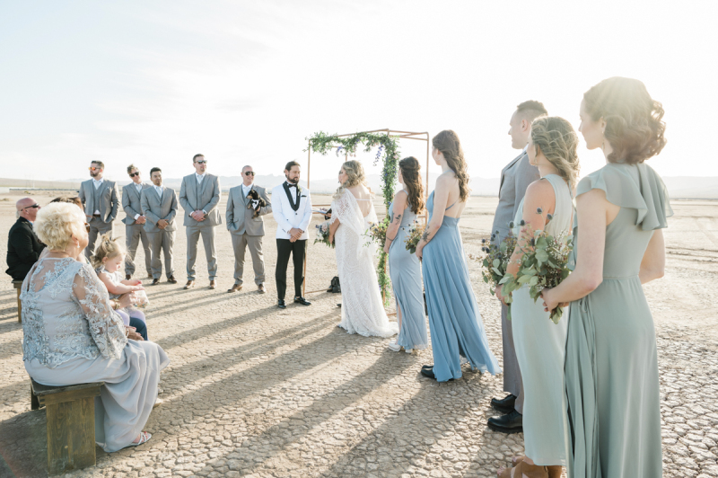A wedding is being held on the Eldorado Dry Lake Bed. The bride and groom are in the middle and are flanked by their wedding party. Guests are seen sitting on wooden benches. A wedding is being held on the Eldorado Dry Lake Bed. The bride and groom are in the middle and are flanked by their wedding party. Guests are seen sitting on wooden benches.