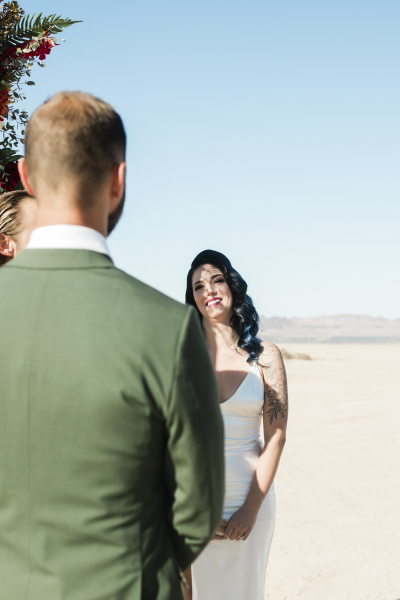 A bride stands with her groom. The photo is taken from behind the groom with the bride facing the camera and smiling at the groom. They are getting married on the Eldorado Dry Lake Bed. A bride stands with her groom. The photo is taken from behind the groom with the bride facing the camera and smiling at the groom. They are getting married on the Eldorado Dry Lake Bed.