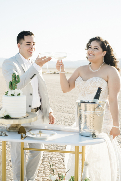 A groom and a bride in a strapless white wedding dress toast each other with Champagne coupe glasses. A wedding cake and an ice bucket with a bottle sit on a table which they stand behind. They are celebrating their wedding on the Dry Lake Bed. A groom and a bride in a strapless white wedding dress toast each other with Champagne coupe glasses. A wedding cake and an ice bucket with a bottle sit on a table which they stand behind. They are celebrating their wedding on the Dry Lake Bed.