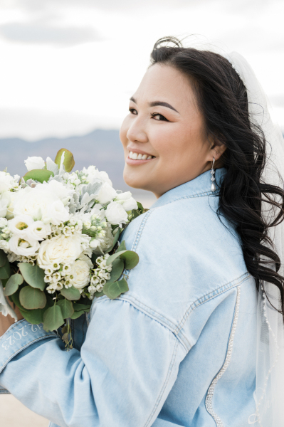A close up of a bride posing for wedding photos. She is holding her flowers about shoulder high as she turns her head back to the left and towards the camera. She is wearing a light blue denim jacket and a white veil. A close up of a bride posing for wedding photos. She is holding her flowers about shoulder high as she turns her head back to the left and towards the camera. She is wearing a light blue denim jacket and a white veil.