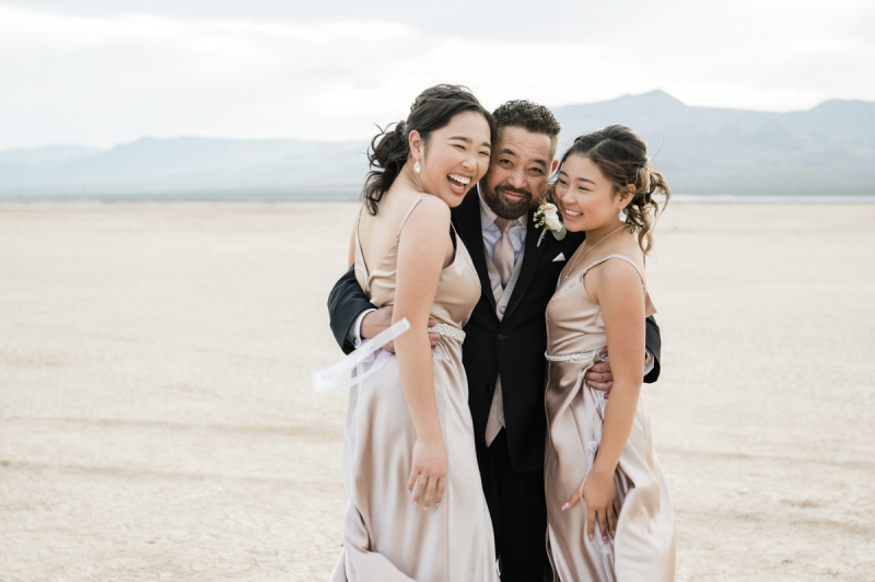 On the Dry Lake Bed outside of Las Vegas a man in a dark suit embraces two women in matching bridesmaids' dresses. The group smiles with delight.