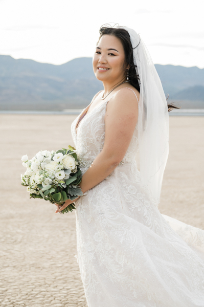 A bride poses on her wedding day at the Eldorado Dry Lake Bed. She is standing at a three-quarters angle from the camera. Mountains can be seen in the distance. A bride poses on her wedding day at the Eldorado Dry Lake Bed. She is standing at a three-quarters angle from the camera. Mountains can be seen in the distance.