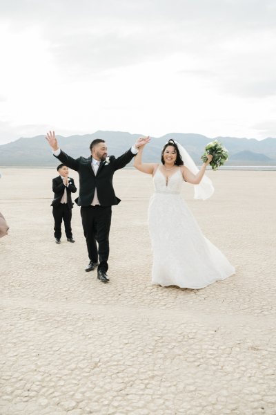 A just married couple raises their hands in the air to celebrate their wedding. They walk on the dry cracked ground of the Dry Lake Bed, while a young boy claps for them in the background. A just married couple raises their hands in the air to celebrate their wedding. They walk on the dry cracked ground of the Dry Lake Bed, while a young boy claps for them in the background.