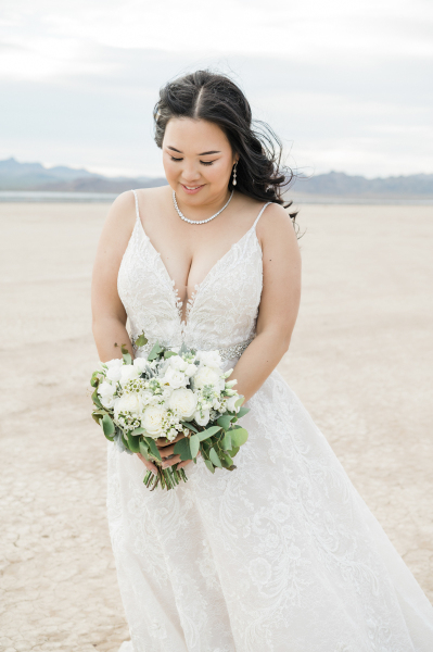 A bride admires her bouquet of white flowers that she holds in front of her. She wears a long sleeveless lace wedding dress with a necklace and earings for her wedding on the Dry Lake Bed in Las Vegas.