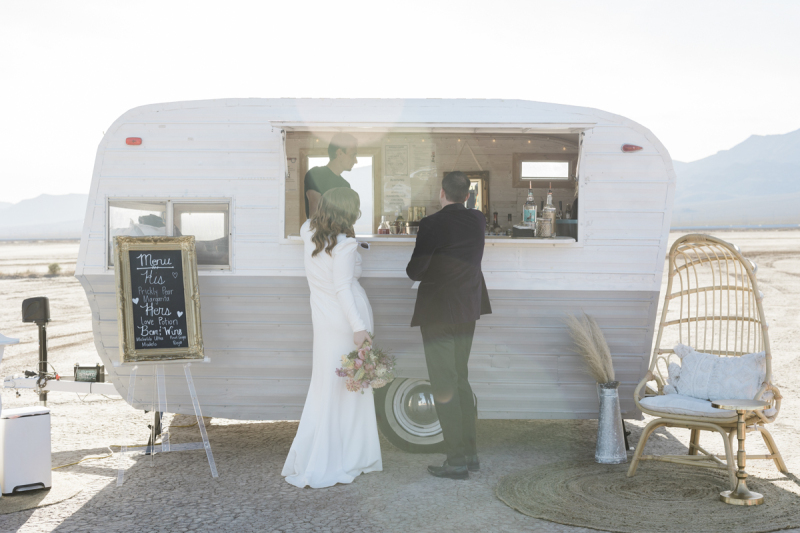 A bride and groom order drinks from a bar that has been built into a vintage camping trailer. Their wedding took place on the Dry Lake Bed south of Las Vegas.