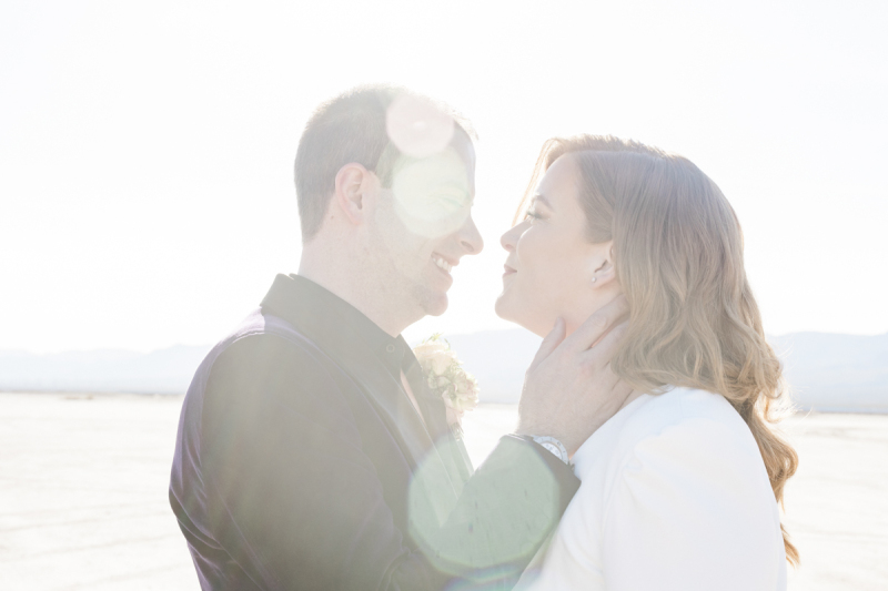 Lens flares partially obscure the face of a groom as he looks lovingly into his bride's eyes and holds her head in his hands. The two look lovingly at each other during their wedding at the Dry Lake Bed.