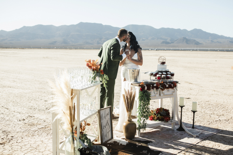 A groom and bride kiss as they celebrate their wedding with cupcakes and Champagne. They are standing behind a pair of small tables arranged with the treats, flowers candles and a sign that reads, "All of me loves all of you". A groom and bride kiss as they celebrate their wedding with cupcakes and Champagne. They are standing behind a pair of small tables arranged with the treats, flowers candles and a sign that reads, "All of me loves all of you".