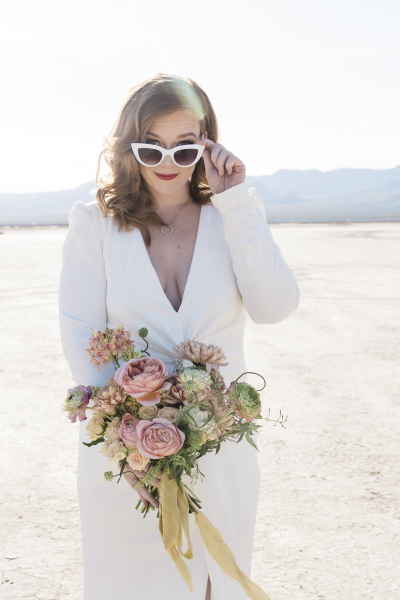 A blond haired woman in a white dress with long sleeves poses for a photo on the Dry Lake Bed. She holds her bouquet of flowers in front of her body with her right hand and tips her large white sunglasses away from her eyes with her left hand.