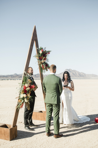 A bride reads her vows to her new husband during their wedding ceremony at the Dry Lake Bed outside of Las Vegas. They are standing at a triangular wooden arbor adorned with flowers. A male officiant stands between the couple.