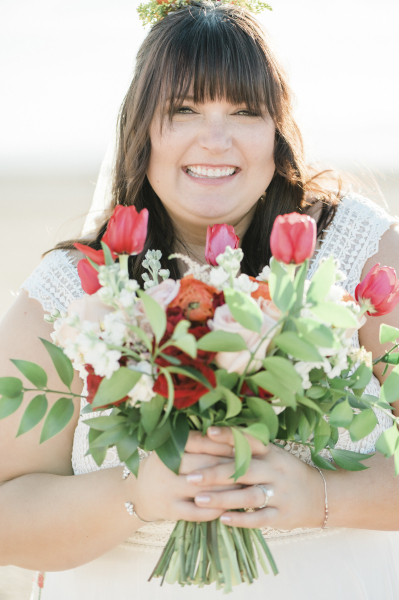 A bride with a big smile holds a bouquet of flowers in front of her body in this close up photo. The bouquet is adorned with red tulips and roses. The woman has long dark hair with bangs covering her eyebrows. A bride with a big smile holds a bouquet of flowers in front of her body in this close up photo. The bouquet is adorned with red tulips and roses. The woman has long dark hair with bangs covering her eyebrows.