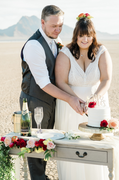 At the Eldorado Dry Lake Bed, a groom and bride cut into a wedding cake. They are standing behind a mini celebration table decorated with flowers that also has a bottle of Champagne, and two Champagne flutes.
