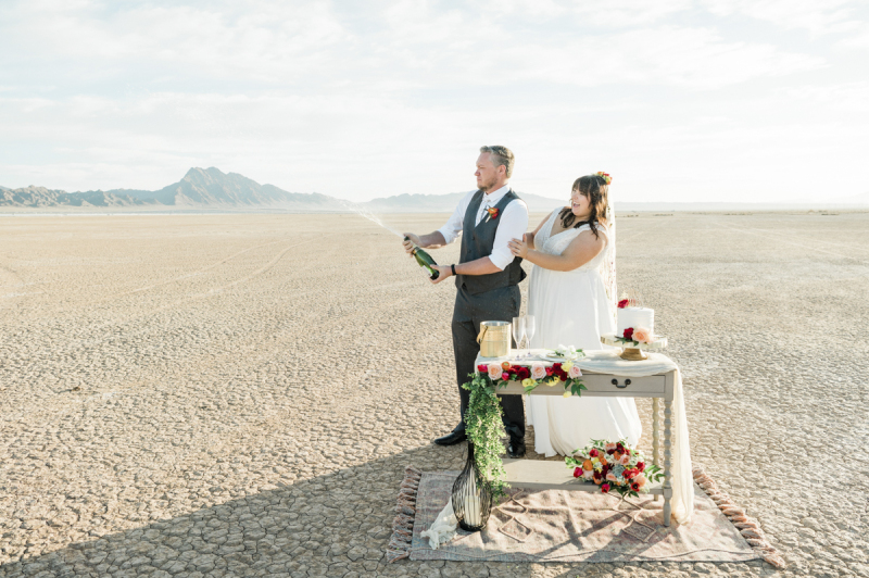 A groom sprays a bottle of Champagne into the air in celebration as his new wife looks on and laughs. The couple is celebrating their wedding on the Dry Lake Bed just south of Boulder City, near Las Vegas, Nevada.