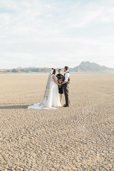 A wide shot of a bride, groom and officiant standing in the middle of the Dry Lake Bed during a wedding ceremony. They are surrounded by nothingness except for the dry cracked ground stretching off into the distance where mountains rise in the background. A wide shot of a bride, groom and officiant standing in the middle of the Dry Lake Bed during a wedding ceremony. They are surrounded by nothingness except for the dry cracked ground stretching off into the distance where mountains rise in the background.