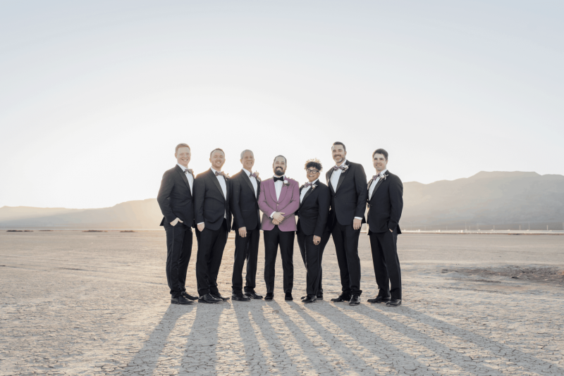 A groom poses at the center of a group photo with his groomsmen. They are standing facing away from the sun and they cast long shadows in front of them onto the ground of the Dry Lake Bed outside of Las Vegas.