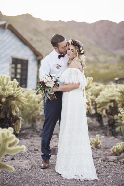 A groom embraces a bride who wears a floral crown as they pose for wedding photos amongst the cacti.