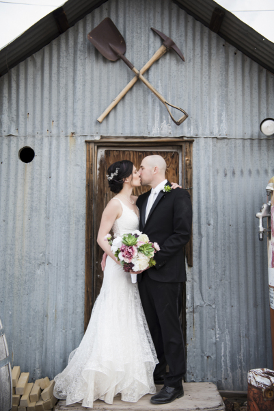 A bride and groom kissing in the doorway of a rustic tin shed with mining tools hung above the door.