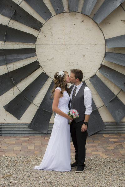 A bride and groom kiss in front of a large vintage windmill blade hanging on a wall.