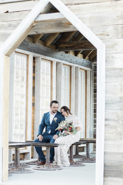 A groom and bride sit on a bench and are viewed through the door of a rustic wooden chapel.