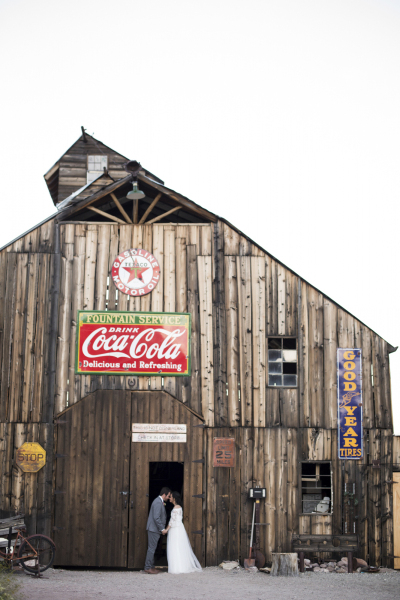 A wide shot of a vintage wooden barn with a groom and bride kissing in the doorway.