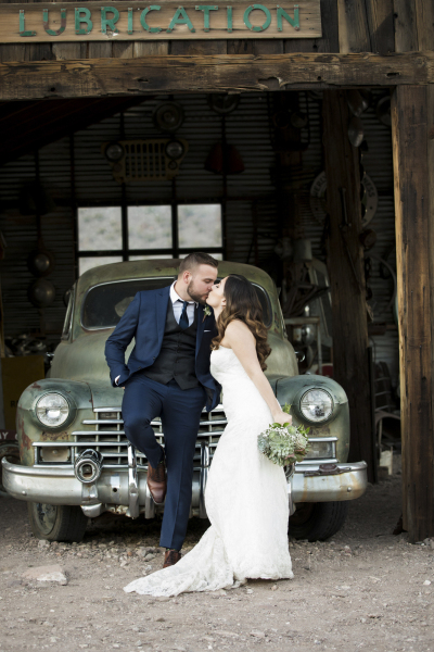 A bride and groom kissing as he leans on the hood of a vintage car in a barn in Eldorado Canyon.