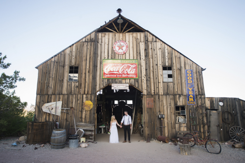 Wide shot of a rustic barn covered in vintage advertising signs. A bride and groom hold hands in the doorway.