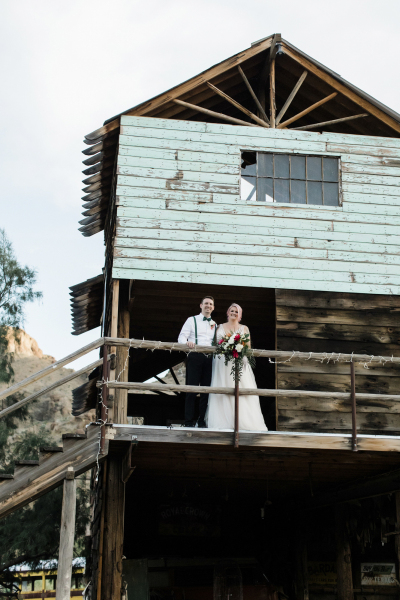 A groom and bride stand on the second-floor balcony that wraps around a vintage wooden structure in Eldorado Canyon.