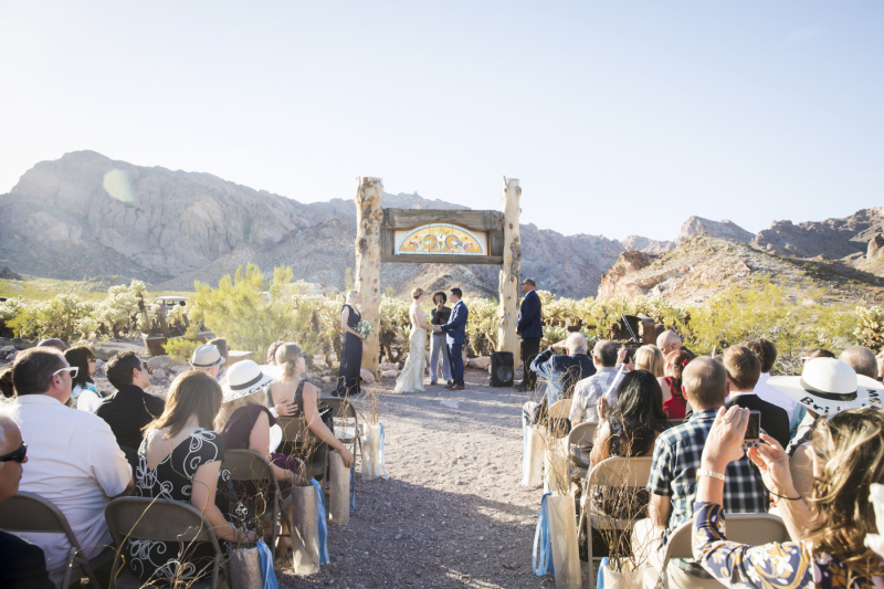 A wedding takes place underneath a rustic stained-glass archway during an outdoor desert wedding.