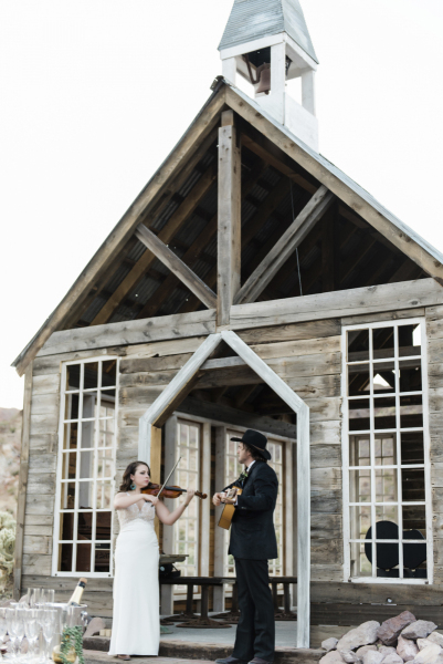 A bride and groom play instruments, her a fiddle and he an acoustic guitar, as they stand on the steps of a rustic wooden chapel.
