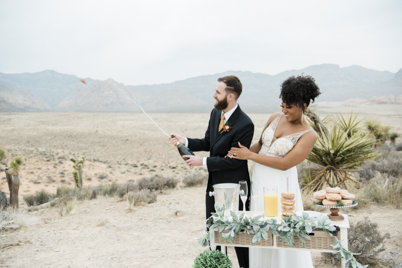 Bride standing behind groom who is popping champagne.