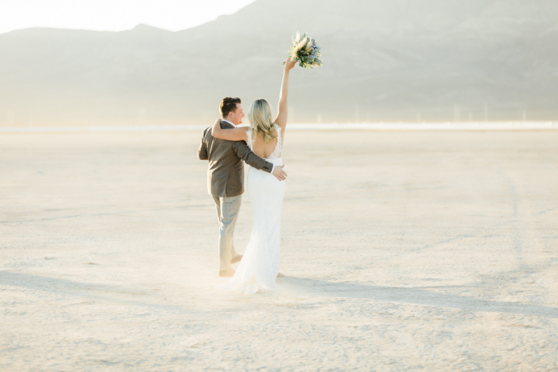 Bride holding wedding bouquet of flowers in air while walking with groom across the dry lake bed.