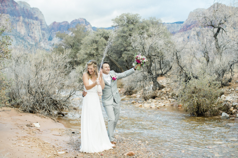 Groom holding bouquet of flowers and bride opening champagne bottle.