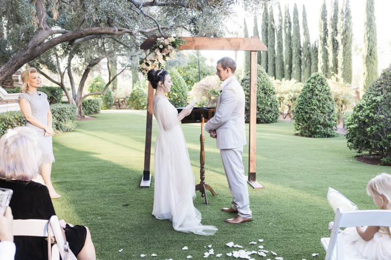 A woman reads vows to her husband as they stand in front of a simple wooden arbor and podium amongst a Cypress tree-lined lawn. Guests are seen in the foreground sitting on white chairs.