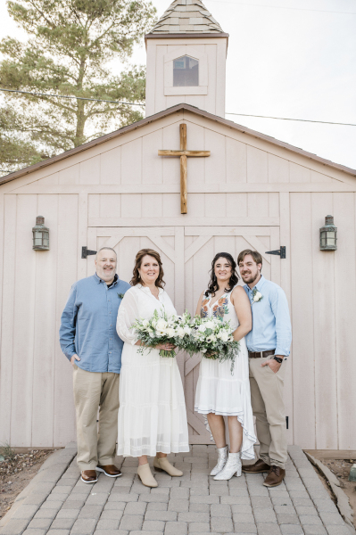 An older husband and wife, and a younger husband and wife stand in front of a rustic wooden chapel after renewing their vows. The women each wear white dresses and hold matching bouquets while the men wear khaki pants and blue dress shirts.