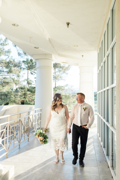A middle-aged couple smile at each other as they walk hand-in-hand on a classical looking veranda. She wears a simple white wedding dress while he wears dark slacks and a dress shirt with a tie.