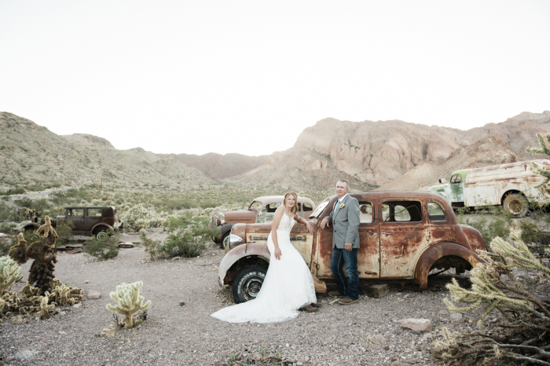 A bride wearing a long white dress sits on the fender of a rusty vintage car while her husband leans against the driver's side window. The car is 1 of 4 pictured in the landscape of a ghost town near Las Vegas.
