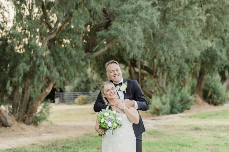 A man stands behind his wife smile and embraces her by the shoulders on the day of their vow renewal ceremony. The backdrop is a row of green trees and grass.
