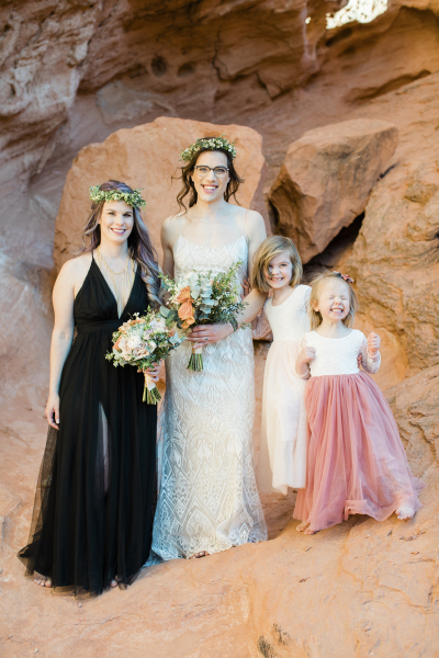 Two women wearing floral crowns and holding bouquets are joined by two little girls on their vow renewal day. The group stands amongst beautifully wind-carved red sandstone.