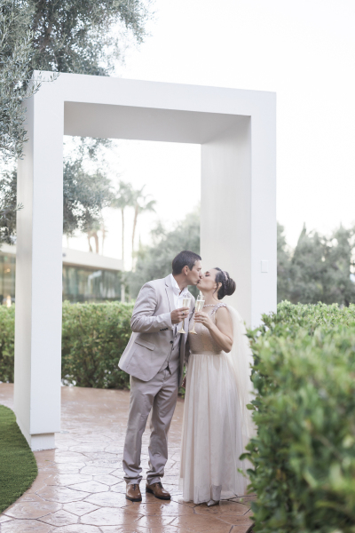 A groom and bride kiss following their vow renewal ceremony. The stamped concrete path they are standing on winds through the trees and bushes underneath a tall rectangular archway.