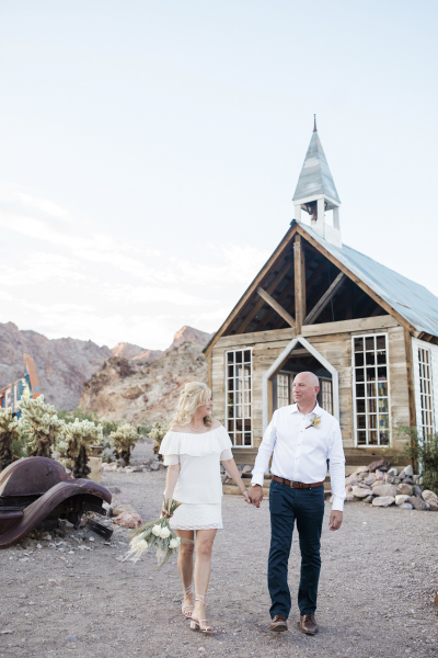 In front of a rustic wooden chapel in the Eldorado Canyon a couple walks hand-in-hand and gazes at each other after renewing their vows with Cactus Collective Weddings.