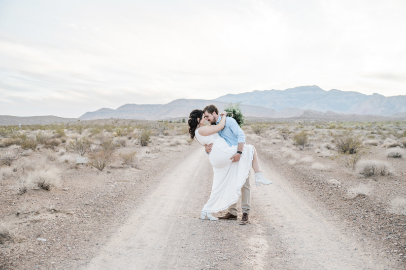A husband lifts his wife's leg and tips her backwards as he pulls her close. They are standing in the middle of a dirt road that leads through the Mojave Desert to some mountains in the distance.