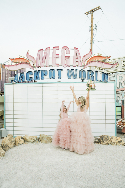 The non-operational marquee for Mega Jackpot World on display at the Neon Museum in Las Vegas serves as a unique backdrop for a woman and her young daughter in matching pink princess dresses. They raise their arms in celebration of the woman's vow renewal day.