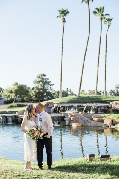 A woman holds a bouquet of flowers while kissing her husband. The couple stands in front a series of cascading waterfalls that flow into a pond with 4 tall palm trees behind the waterfalls.