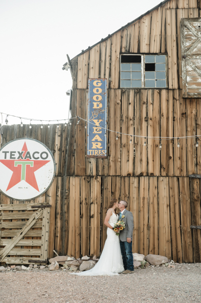 A wide shot of a bride and groom kissing in front of the outside wall of a faded old barn. The barn is decorated with a string of bistro lights, an old Texaco gas station sign and an old Goodyear Tires sign.
