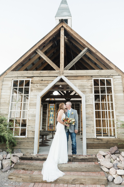 A bride and groom kiss standing on the stairs in front of a rustic western chapel. There are large colonial windows on the front of the building, and through the open door you can see a stained-glass window at the back of the building.