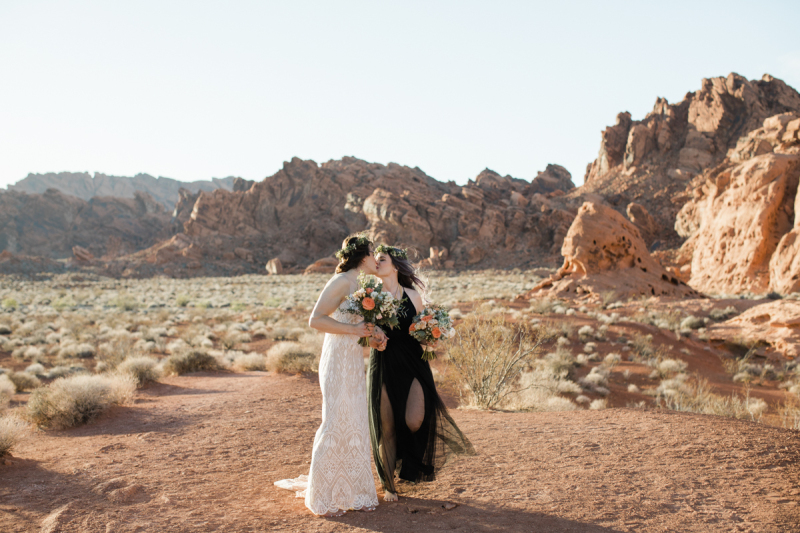 A woman in a white lacy dress and floral crown kisses her wife who is wearing a black dress and a matching floral crown. They are surrounded by dramatic red sandstone rock outcroppings and hundreds of small desert bushes.