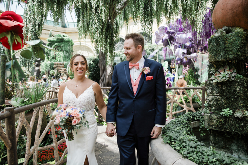 A bride and groom hold hands and walk through the conservatory at the Bellagio Hotel and Casino in Las Vegas. They are surrounded by greenery, a fountain covered in succulents, oversized red and purple flowers and a wooden railing. The groom's pink vest, bow tie and pocket square match the flowers in the bride's bouquet.