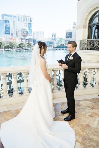 A bride and groom are getting married on a balcony overlooking the lake at the Bellagio Hotel and Casino in Las Vegas. The groom reads his vows from a little black notebook. Other buildings on the Strip are visible across the lake in the background.