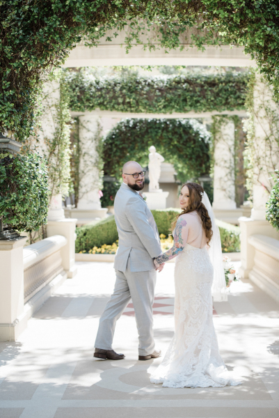 A groom and bride pose underneath a series of formal garden archways on their wedding day. They face away from the camera while turning their heads and shoulders inwards towards each other and back towards the camera.
